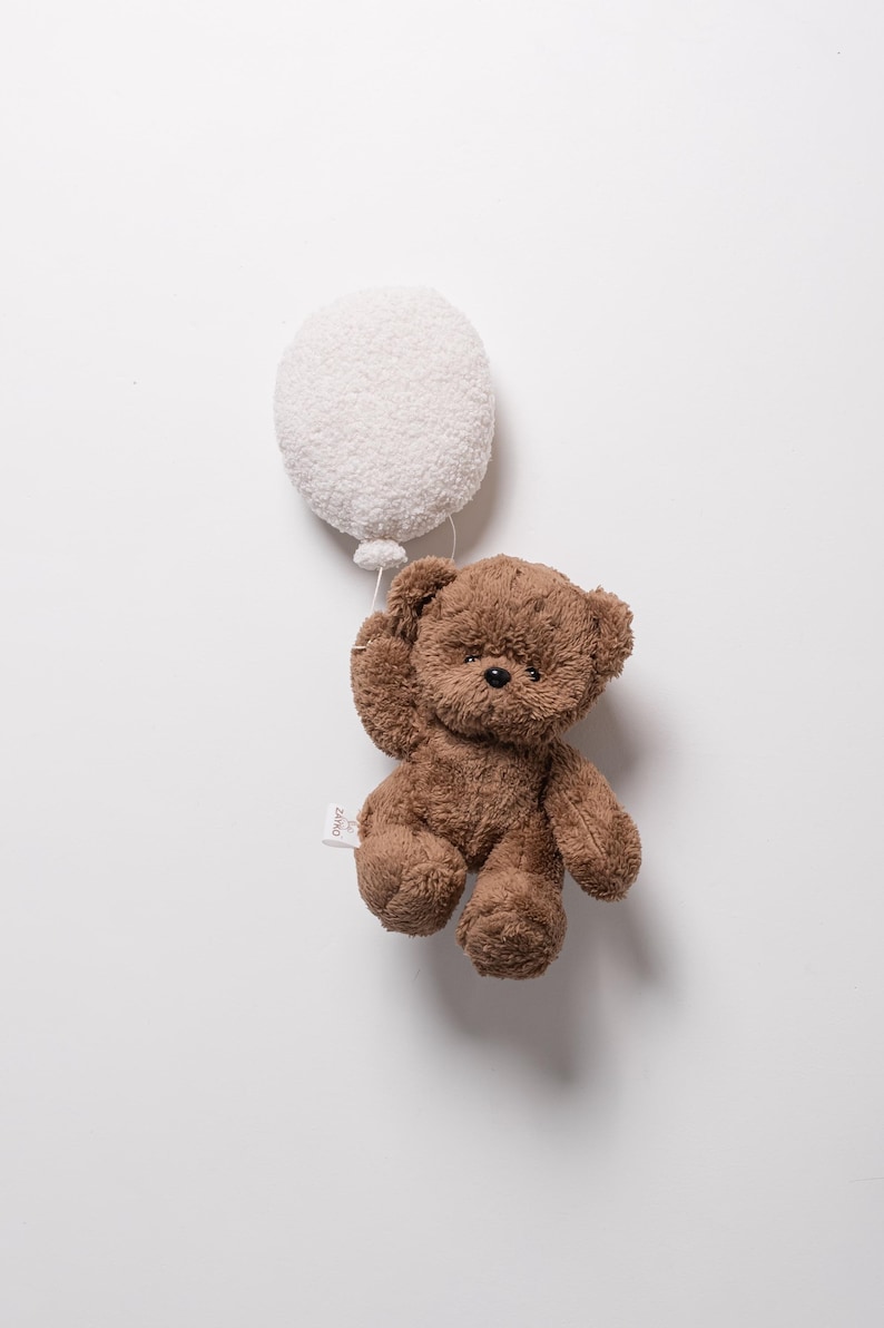Pode incluir: Um urso de pel&uacute;cia marrom segurando um bal&atilde;o branco e fofo. O urso tem uma pequena etiqueta anexada. O brinquedo est&aacute; em um fundo branco liso.