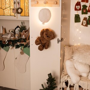 Pode incluir: Um cabide de parede de madeira com detalhes de orelhas de coelho e tr&ecirc;s pinos. Uma luz de bal&atilde;o branco brilhante e um urso de pel&uacute;cia marrom est&atilde;o presos &agrave; porta. Decora&ccedil;&otilde;es de Natal no fundo.