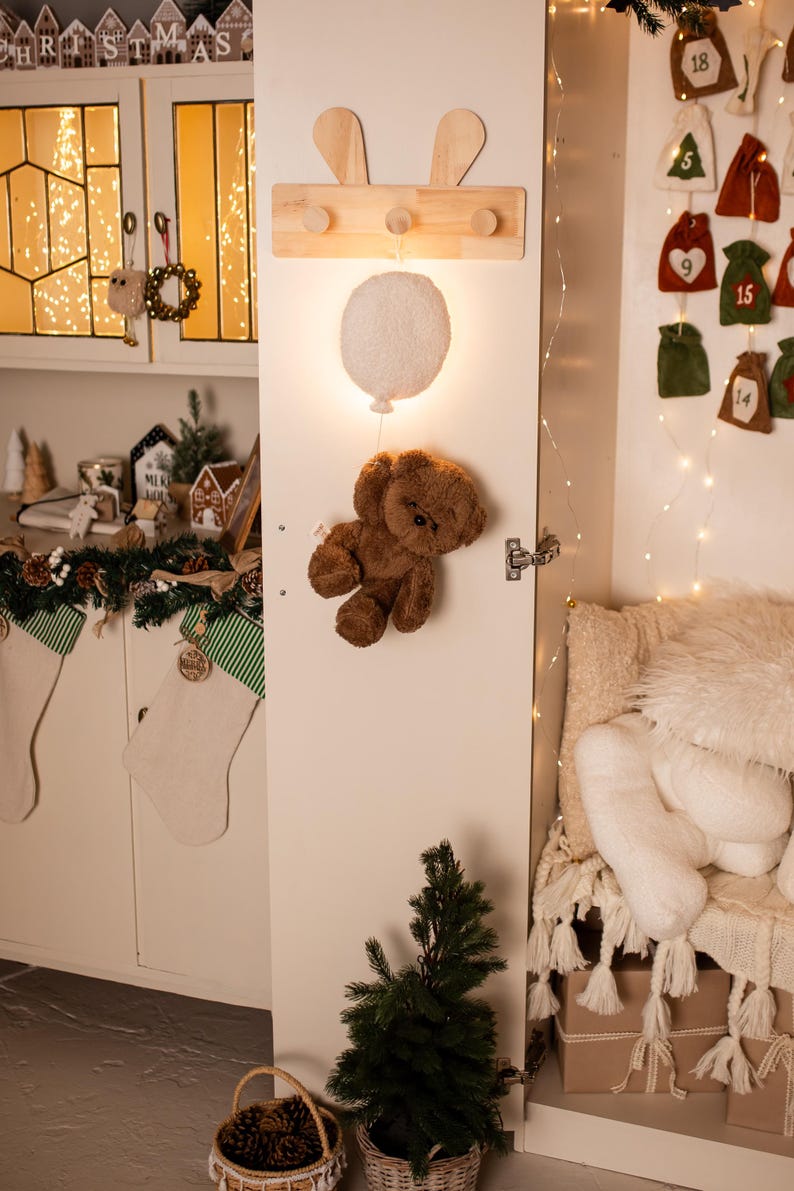 Pode incluir: Um cabide de parede de madeira com detalhes de orelhas de coelho e tr&ecirc;s pinos. Uma luz de bal&atilde;o branco brilhante e um urso de pel&uacute;cia marrom est&atilde;o presos &agrave; porta. Decora&ccedil;&otilde;es de Natal no fundo.