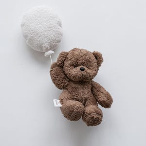 Pode incluir: Um urso de pel&uacute;cia marrom segurando um bal&atilde;o branco. O urso tem uma pequena etiqueta presa ao corpo. O bal&atilde;o &eacute; redondo e fofo, e o urso parece estar flutuando.
