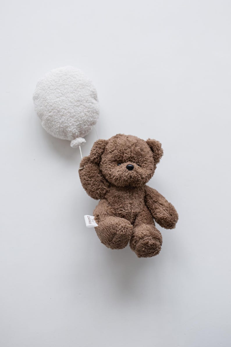Pode incluir: Um urso de pel&uacute;cia marrom segurando um bal&atilde;o branco. O urso tem uma pequena etiqueta presa ao corpo. O bal&atilde;o &eacute; redondo e fofo, e o urso parece estar flutuando.