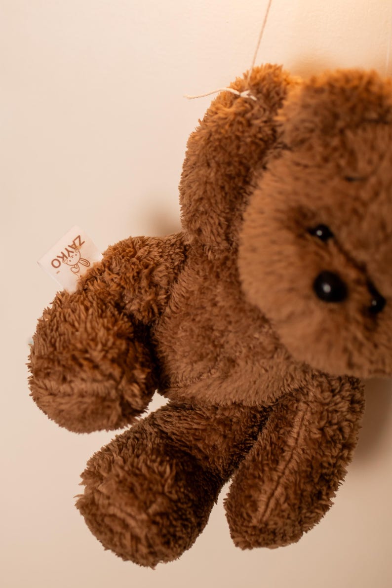 Pode incluir: Um urso de pel&uacute;cia marrom com uma textura macia e fofa. O urso tem olhos pretos e uma pequena etiqueta que diz "ZAYKO". O brinquedo de pel&uacute;cia est&aacute; pendurado por um fio, com seus bra&ccedil;os e pernas vis&iacute;veis.