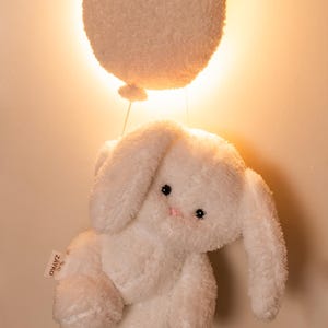 Pode incluir: Um coelho de pel&uacute;cia branco com orelhas compridas e um nariz rosa, suspenso sob um bal&atilde;o branco, redondo e fofo. O coelho tem olhos pretos e est&aacute; preso ao bal&atilde;o por um fio fino. O fundo &eacute; uma luz quente e suave.