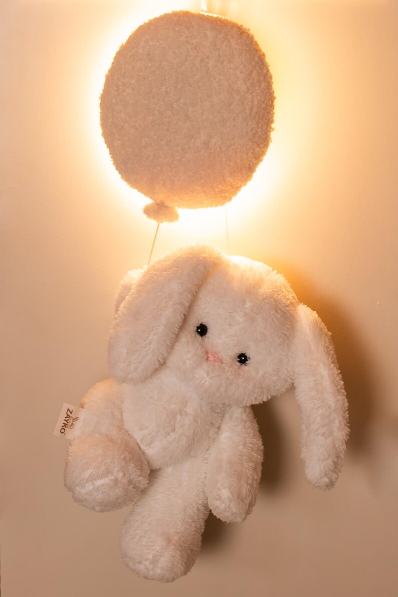 Pode incluir: Um coelho de pel&uacute;cia branco com orelhas compridas e um nariz rosa, suspenso sob um bal&atilde;o branco, redondo e fofo. O coelho tem olhos pretos e est&aacute; preso ao bal&atilde;o por um fio fino. O fundo &eacute; uma luz quente e suave.