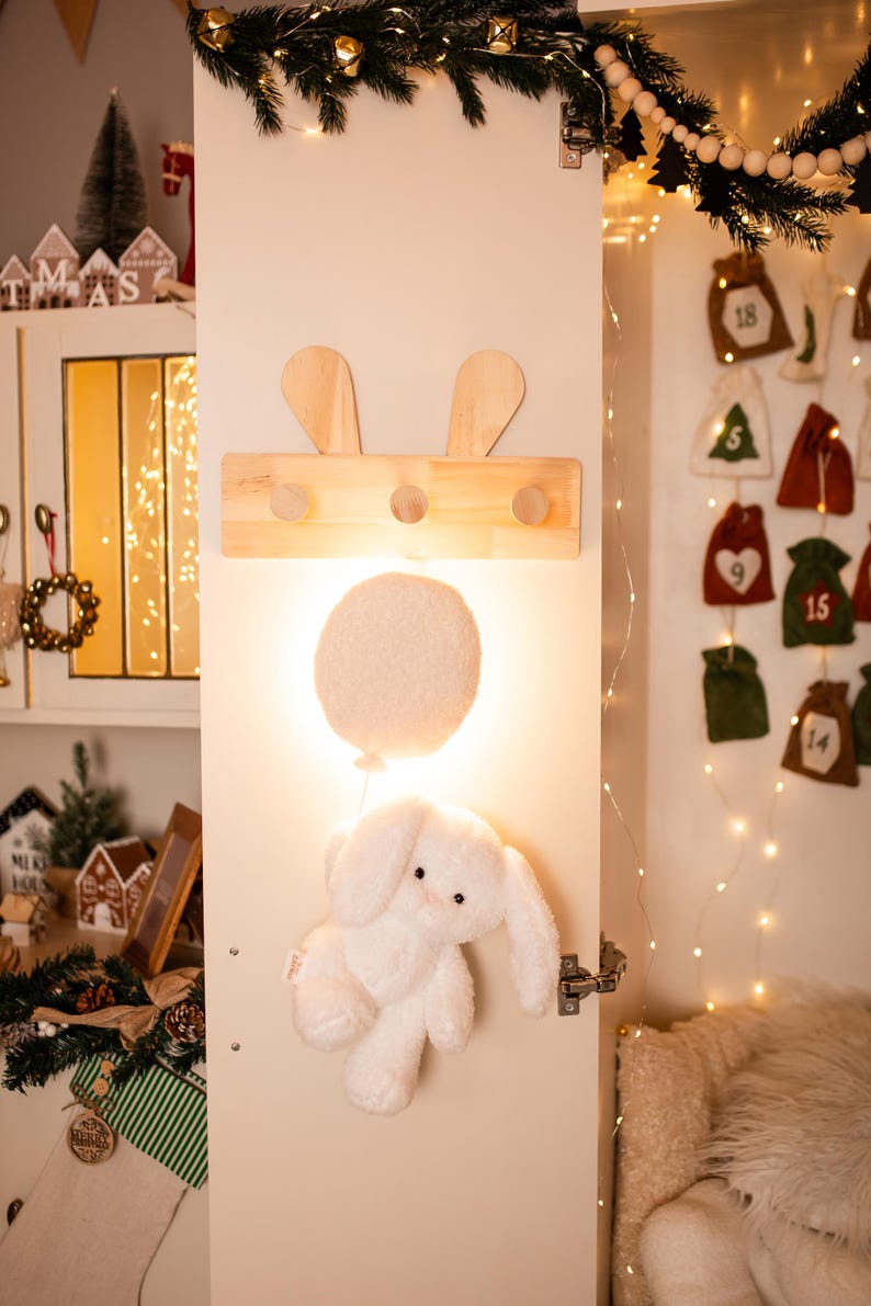Pode incluir: Um cabide de madeira com detalhes de orelhas de coelho e tr&ecirc;s pinos, montado em uma porta branca. Abaixo, um coelho de pel&uacute;cia branco e fofo pendura, com uma luz em forma de bal&atilde;o brilhante. Decora&ccedil;&otilde;es de f&eacute;rias.