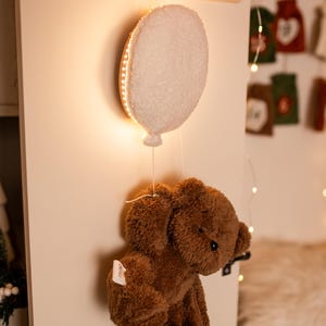 Pode incluir: Uma decora&ccedil;&atilde;o de ber&ccedil;&aacute;rio extravagante com um urso de pel&uacute;cia castanho suspenso de uma lumin&aacute;ria em forma de bal&atilde;o branco. A luz &eacute; delineada com luzes LED quentes, criando um brilho suave. A lumin&aacute;ria &eacute; montada em uma parede de madeira com um cabide.