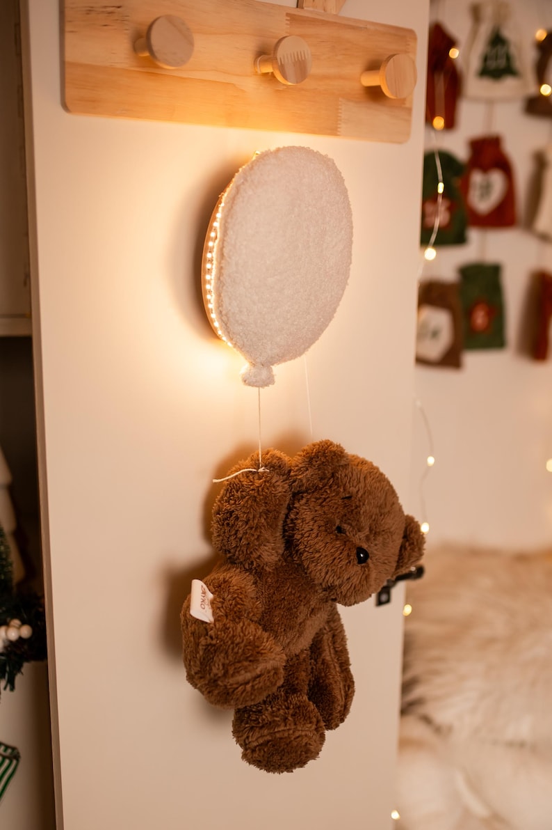 Pode incluir: Uma decora&ccedil;&atilde;o de ber&ccedil;&aacute;rio extravagante com um urso de pel&uacute;cia castanho suspenso de uma lumin&aacute;ria em forma de bal&atilde;o branco. A luz &eacute; delineada com luzes LED quentes, criando um brilho suave. A lumin&aacute;ria &eacute; montada em uma parede de madeira com um cabide.
