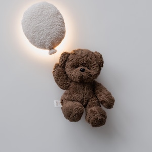 Pode incluir: Um urso de pel&uacute;cia castanho com uma etiqueta, posicionado ao lado de uma lumin&aacute;ria branca em forma de bal&atilde;o. O urso tem um pequeno nariz preto e levanta o bra&ccedil;o. O fundo &eacute; uma parede branca lisa.