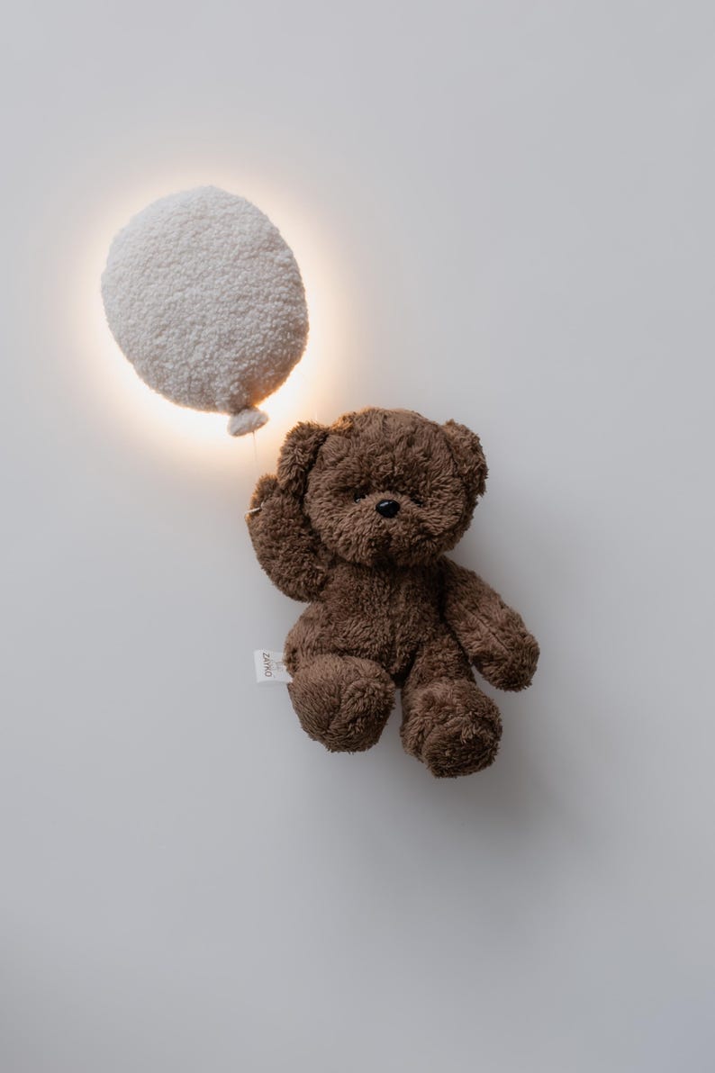 Pode incluir: Um urso de pel&uacute;cia castanho com uma etiqueta, posicionado ao lado de uma lumin&aacute;ria branca em forma de bal&atilde;o. O urso tem um pequeno nariz preto e levanta o bra&ccedil;o. O fundo &eacute; uma parede branca lisa.