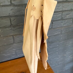 May include: A light beige leather tote bag with a long strap and a flap closure. The bag has a raw edge and is made of a soft, supple leather.