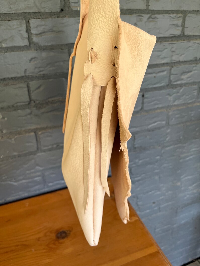 May include: A light beige leather tote bag with a long strap and a flap closure. The bag has a raw edge and is made of a soft, supple leather.