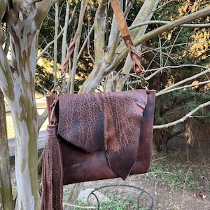 May include: A brown leather crossbody bag with a flap closure and a long, adjustable strap. The bag features a decorative tassel and is suspended from a tree branch. The leather has a distressed appearance, adding to its rustic charm.