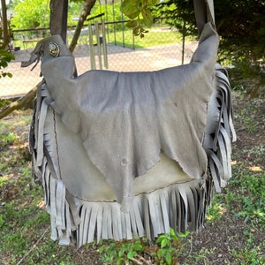 May include: A gray leather shoulder bag with fringe detailing. The bag has a flap closure and a silver button.