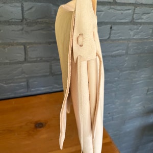 May include: A cream-colored leather satchel bag with a long strap. The bag is folded over and has a flap closure. The strap is attached to the bag with a metal buckle.