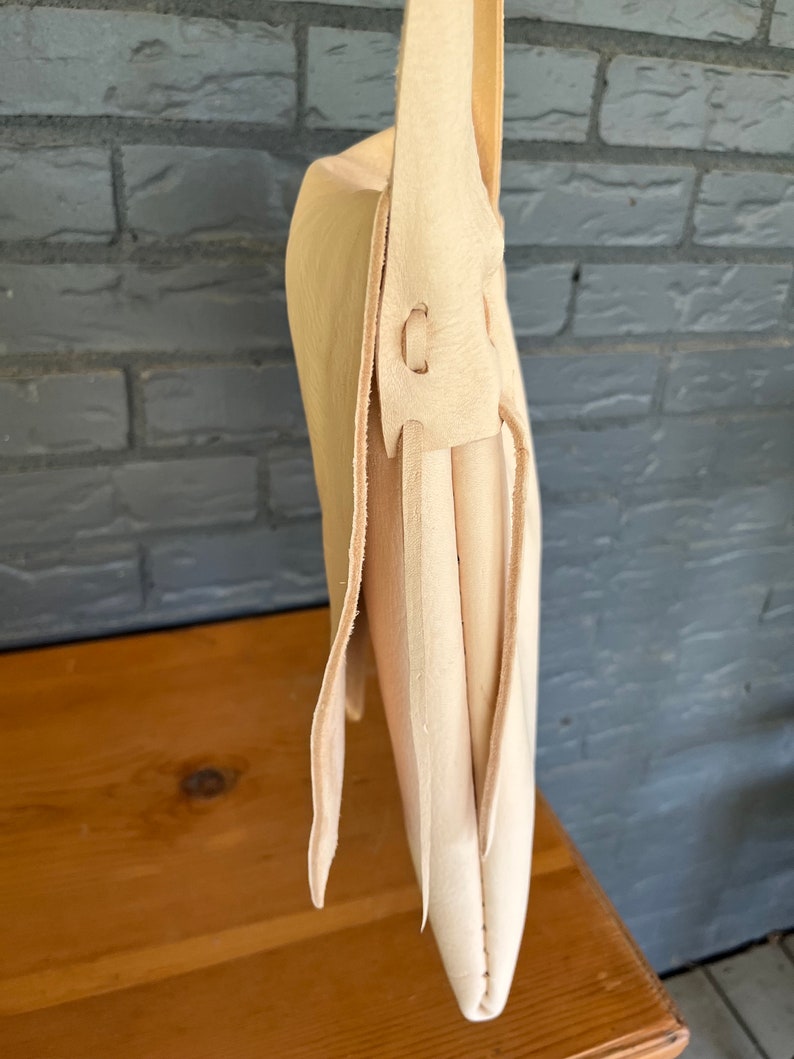 May include: A cream-colored leather satchel bag with a long strap. The bag is folded over and has a flap closure. The strap is attached to the bag with a metal buckle.