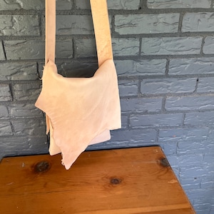 May include: A light brown leather crossbody bag with a long strap. The bag is hanging on a brick wall.