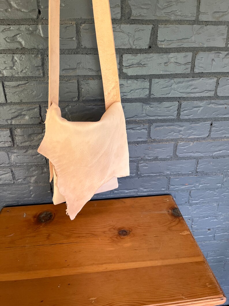 May include: A light brown leather crossbody bag with a long strap. The bag is hanging on a brick wall.