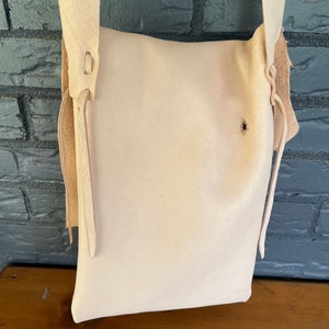 May include: A white leather satchel bag with a long strap and a flap closure. The bag has a simple design and is made of high-quality leather.