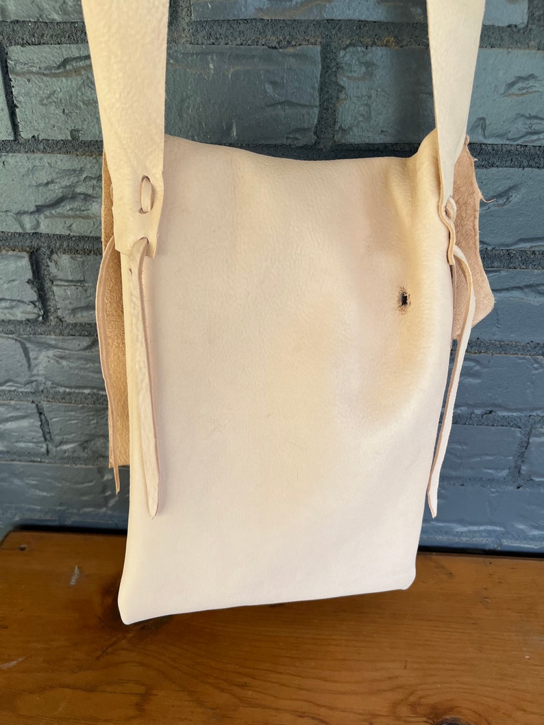May include: A white leather satchel bag with a long strap and a flap closure. The bag has a simple design and is made of high-quality leather.