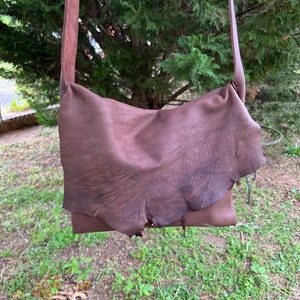 May include: A brown leather crossbody bag with a long strap. The bag has a flap that is folded over and secured with a snap. The bag is made of a soft, supple leather and has a rustic, vintage look.