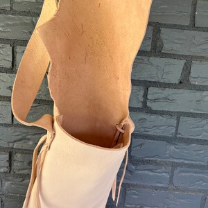 May include: A light brown leather satchel bag with a long strap and a drawstring closure. The bag is empty and hanging from a brick wall.