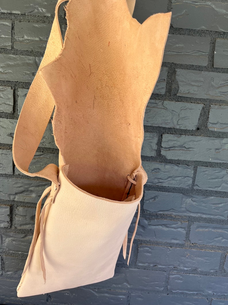 May include: A light brown leather satchel bag with a long strap and a drawstring closure. The bag is empty and hanging from a brick wall.