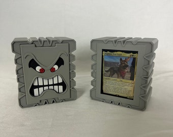 Puede incluir: Dos cajas de cartas impresas en 3D de color gris. Una caja tiene una cara de dibujos animados enojada con ojos rojos y dientes blancos. La otra caja tiene una carta de Magic: The Gathering dentro con el texto "Dogmeat, Ever Loyal" y una imagen de un perro.