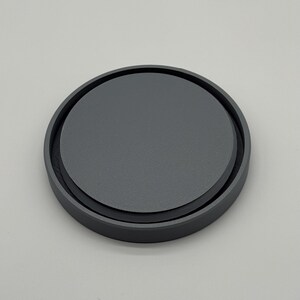 May include: A round, dark gray coaster with a slightly raised edge. The coaster has a matte finish and a simple, minimalist design. The coaster is made of a solid material and appears durable.