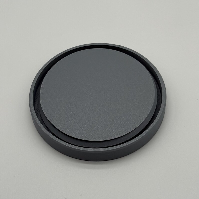 May include: A round, dark gray coaster with a slightly raised edge. The coaster has a matte finish and a simple, minimalist design. The coaster is made of a solid material and appears durable.