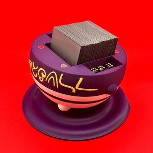 May include: A purple card holder with gold lettering and a white stripe, holding a stack of cards. The holder has a small compartment for dice and sits on a matching purple base. The background is a solid red.