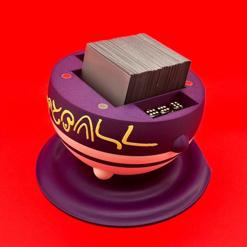 May include: A purple card holder with gold lettering and a white stripe, holding a stack of cards. The holder has a small compartment for dice and sits on a matching purple base. The background is a solid red.