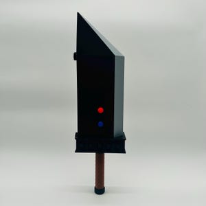 May include: A black, angular sword prop with a brown handle and a black base. The sword features red and blue dot details. The handle has a textured, wrapped appearance. The blade has a distinctive, angled design.