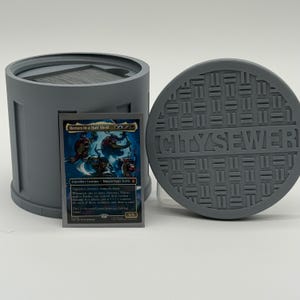 Può includere: Un porta carte grigio cilindrico con un coperchio di tombino abbinato. Una carta collezionabile con il testo "Heroes in a Half Shell" e un'illustrazione di Tartaruga Ninja Mutante è di fronte al supporto. Il coperchio del tombino ha il testo "CITYSEWER".