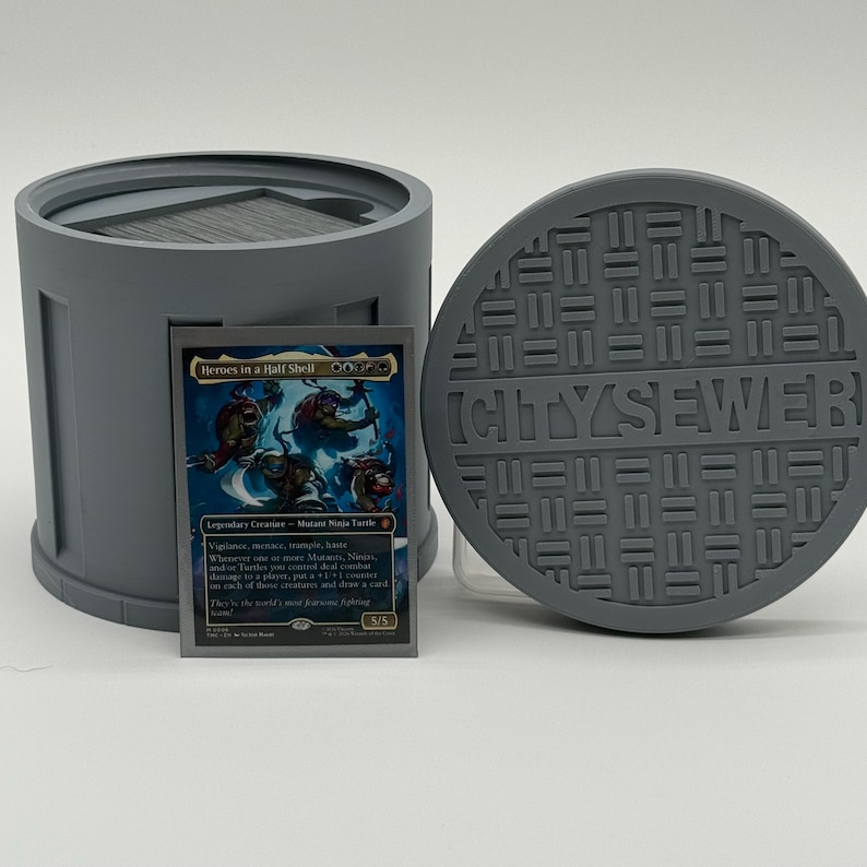 May include: A gray, cylindrical card holder with a matching manhole cover lid. A trading card with the text "Heroes in a Half Shell" and a Mutant Ninja Turtle illustration is in front of the holder. The manhole cover has the text "CITYSEWER".