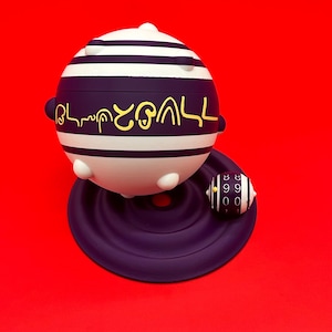 May include: A spherical, white and purple object with horizontal stripes and gold lettering, resting on a purple base. A smaller, striped, cylindrical object with numbers sits nearby. The background is a solid red.