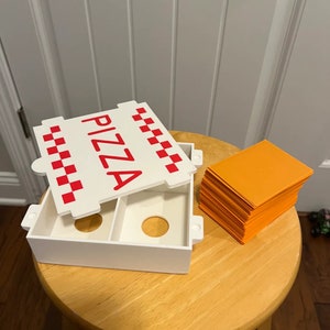 Op de afbeelding: Een witte plastic pizzadoos met een rood-wit geruit patroon en het woord "PIZZA" in rode letters. De doos is open en er ligt een stapel oranje kaarten naast.