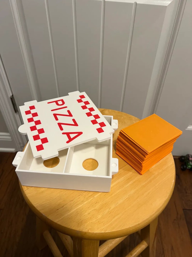Op de afbeelding: Een witte plastic pizzadoos met een rood-wit geruit patroon en het woord "PIZZA" in rode letters. De doos is open en er ligt een stapel oranje kaarten naast.