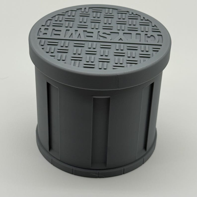 May include: A cylindrical, gray container with a removable lid. The lid features a repeating geometric pattern and text. The container's body has vertical rectangular indentations. The overall design suggests a storage or decorative item.