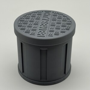 May include: A gray cylindrical object resembling a miniature sewer cover. The top features a grid pattern and the embossed text "CITY SEWER." The sides have vertical rectangular indentations. The object is made of a solid, matte material.
