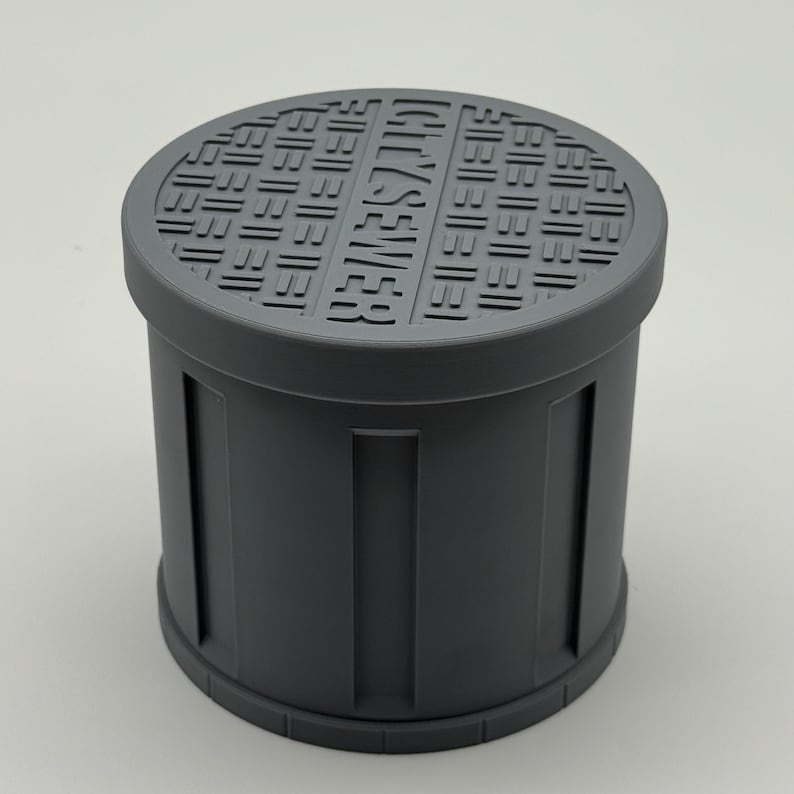 May include: A gray cylindrical object resembling a miniature sewer cover. The top features a grid pattern and the embossed text "CITY SEWER." The sides have vertical rectangular indentations. The object is made of a solid, matte material.