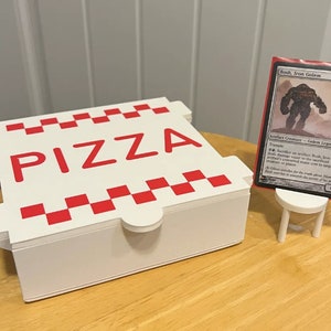 Op de afbeelding: Een witte pizzadoos met een rood-wit geruit patroon en het woord "PIZZA" in rode letters. Een witte standaard houdt een Magic: The Gathering kaart vast met de tekst "Bosh, Iron Golem" en een afbeelding van een grote, grijze golem.