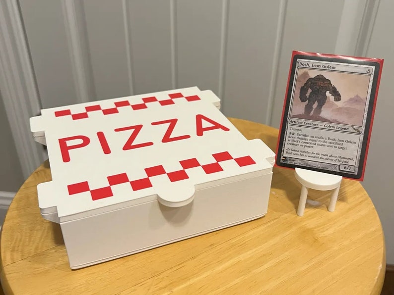 Op de afbeelding: Een witte pizzadoos met een rood-wit geruit patroon en het woord "PIZZA" in rode letters. Een witte standaard houdt een Magic: The Gathering kaart vast met de tekst "Bosh, Iron Golem" en een afbeelding van een grote, grijze golem.