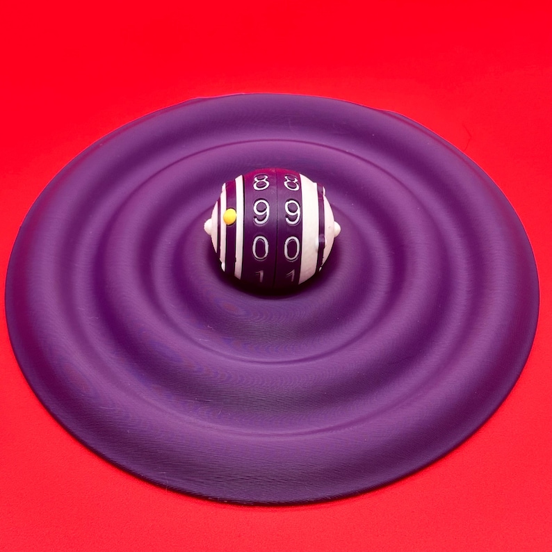May include: A close-up shot of a white and black striped object with numbers on it, resting on a purple, rippled surface. The object has a yellow dot on its side. The background is a solid red.