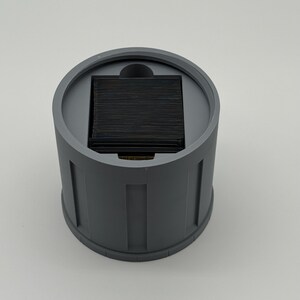 May include: A cylindrical, gray container with a square, black insert. The container has vertical, rectangular indentations around its exterior. The top of the container has a circular rim and a small, curved indentation.