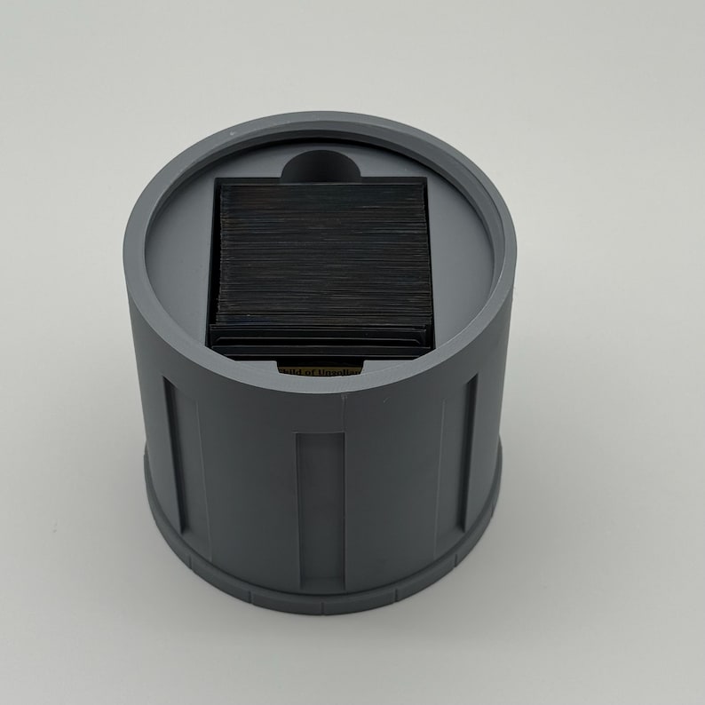 May include: A cylindrical, gray container with a square, black insert. The container has vertical, rectangular indentations around its exterior. The top of the container has a circular rim and a small, curved indentation.