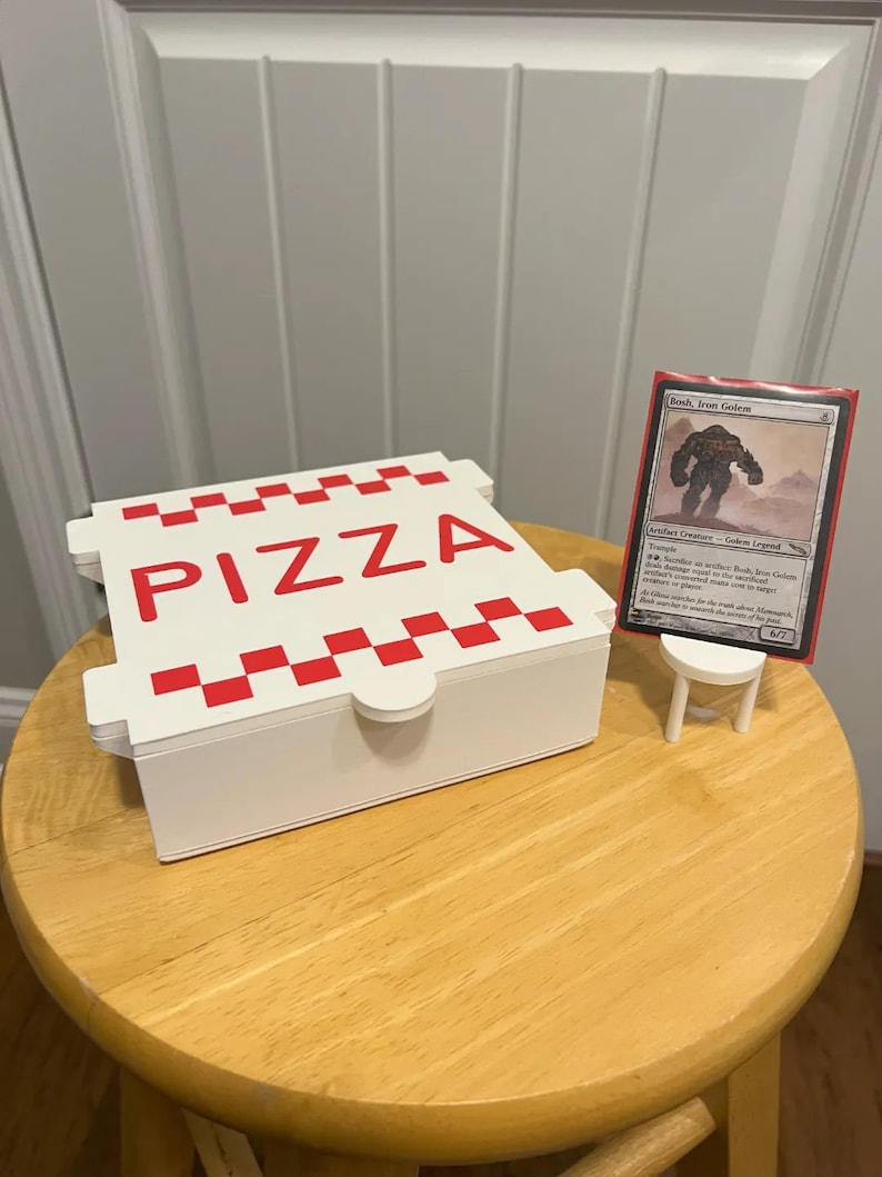 Op de afbeelding: Een witte pizzadoos met een rood-wit geruit patroon en het woord "PIZZA" in rode letters. Een rode en witte Magic: The Gathering kaart met de tekst "Bosh, Iron Golem" wordt getoond op een kleine witte standaard.