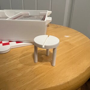 Op de afbeelding: Een witte, miniatuur, houten kruk met een ronde zitting en vier poten. De kruk staat op een houten tafel.