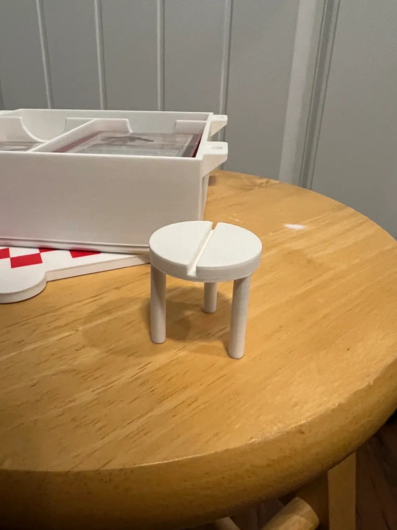 Op de afbeelding: Een witte, miniatuur, houten kruk met een ronde zitting en vier poten. De kruk staat op een houten tafel.
