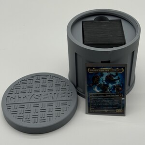 May include: A gray, 3D-printed card holder shaped like a sewer, with a matching lid. The lid reads "CITY SEWER". A trading card with the text "Heroes in a Half Shell" is displayed next to the holder.