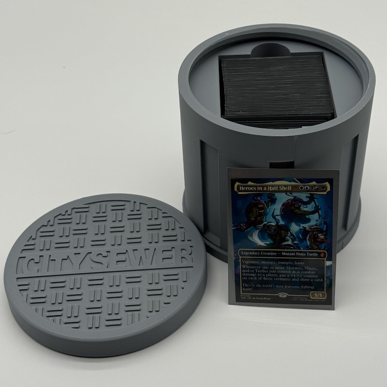 May include: A gray, 3D-printed card holder shaped like a sewer, with a matching lid. The lid reads "CITY SEWER". A trading card with the text "Heroes in a Half Shell" is displayed next to the holder.